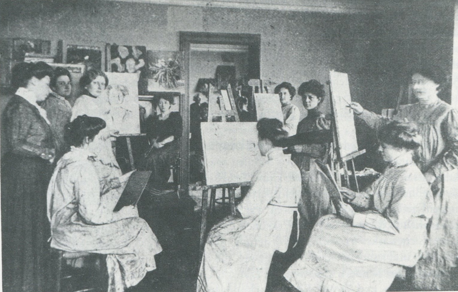 Foto aus der Malschule Gerda Koppel, 1910, Fotograf*in unbekannt Schwarz-weiß Fotografie einer Gruppe von Frauen, die vor Leinwänden sitzen und ein Portrait einer Frau malen, die auf einem Stuhl sitzt.